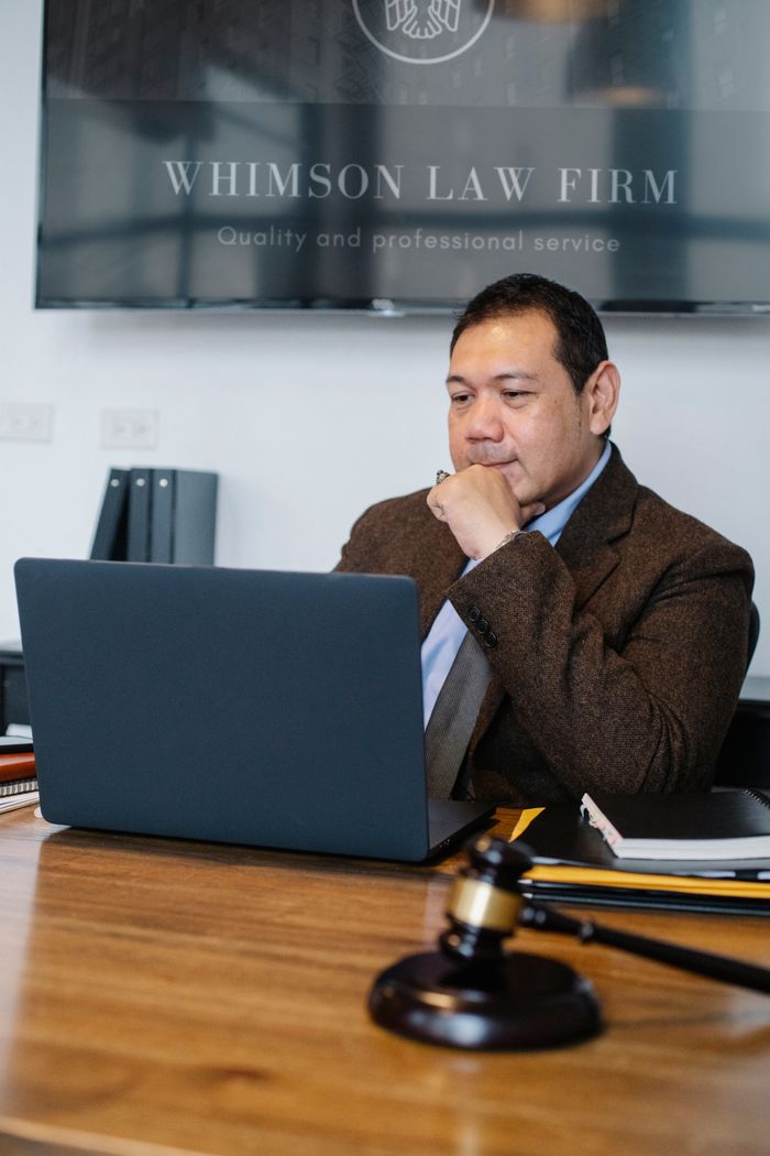 Concentrated Asian male lawyer wearing classy formal suit working on netbook while sitting at table with decorative hammer of justice in modern law office